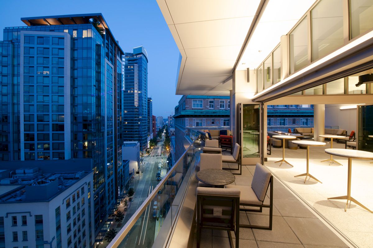 A modern urban balcony view at dusk, overlooking city buildings and a lively street, with outdoor furniture and warm lighting.