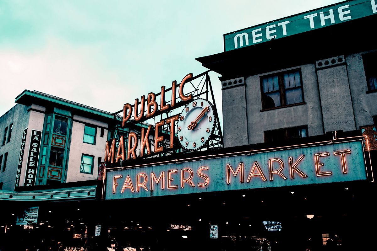 The image shows a public market and farmers market sign with a clock on it, set against an urban backdrop with buildings.