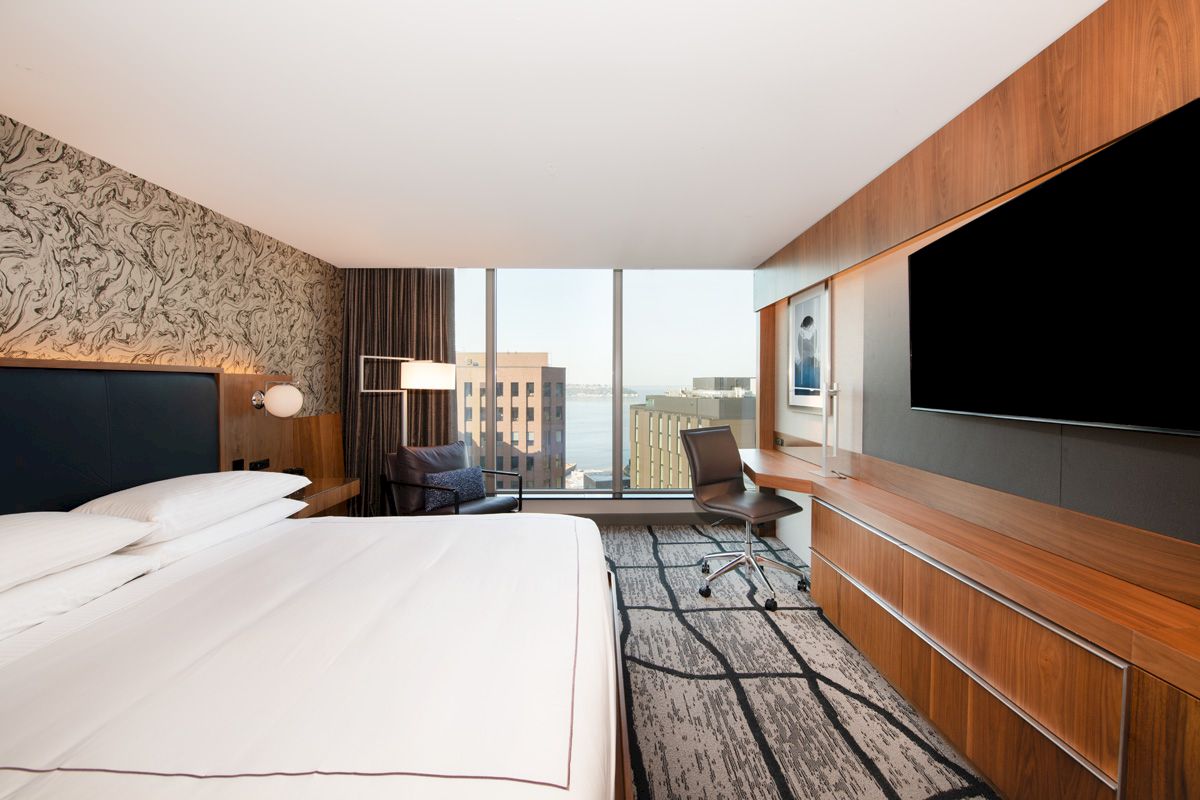 The image shows a modern hotel room with a large bed, desk, chair, wall-mounted TV, and a window view of buildings and water.