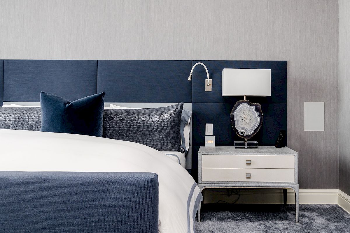 A modern bedroom with a blue and white theme, featuring a bed, nightstand, decorative lamp, and plush carpet flooring.