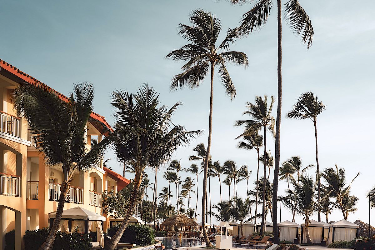 A scenic view of a poolside area with lounge chairs, palm trees, and a resort building under a clear sky.