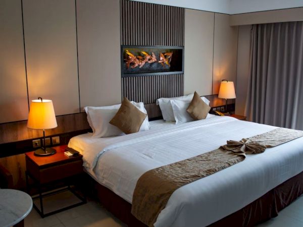 A cozy hotel room with a large bed, decorative pillows, two lamps, and a hanging artwork above. The ambiance is warm and inviting.