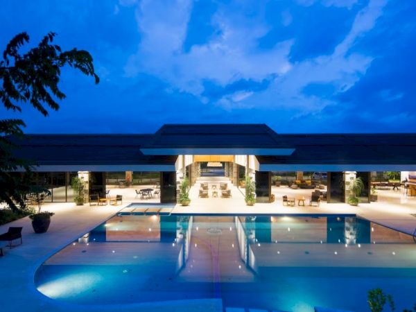 The image shows a luxurious pool area at dusk, featuring a modern building with open seating and a clear, illuminated swimming pool.