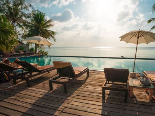 A serene poolside scene with lounge chairs, umbrellas, palm trees, and a calm body of water under a partly cloudy sky.