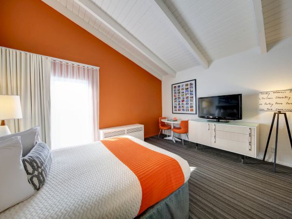 A modern bedroom with an orange accent wall features a bed, TV, desk, chair, and floor lamp, under a vaulted ceiling.