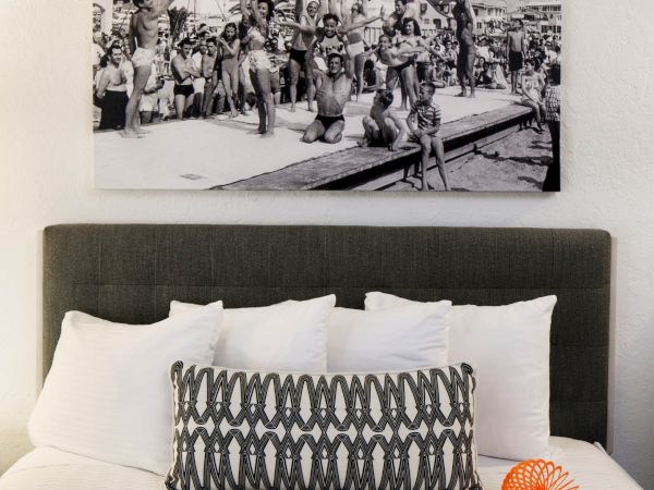 A neatly made bed with white and patterned pillows, an orange object, and a black-and-white photo of people diving by the beach above.