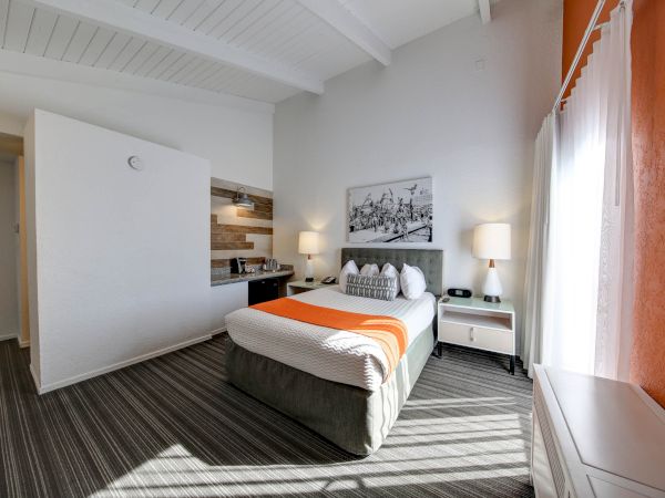The image shows a modern hotel room with a bed, two lamps, and a small kitchenette, featuring a mix of orange, white, and grey accents.