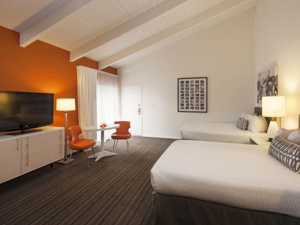 A modern hotel room with two beds, orange and white decor, a TV, and a small dining area with chairs.
