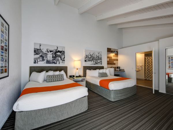 The image shows a hotel room with two beds, overhead beams, orange accents, photographs on the walls, and an open doorway to a bathroom.