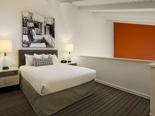 The image shows a modern bedroom with a bed, two lamps, a black-and-white photo on the wall, and a red accent wall in the background.