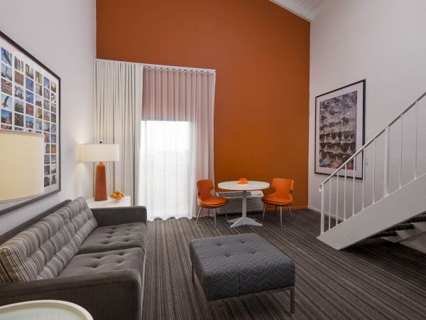 The image shows a modern living room with a gray sofa, orange accent wall, art, a staircase, and a small table with orange chairs.
