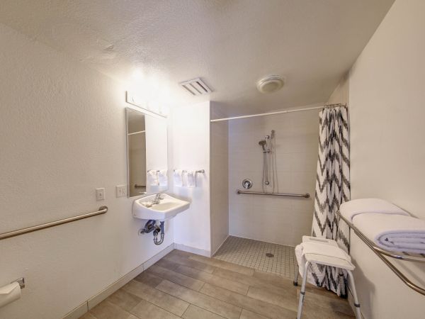 The image shows an accessible bathroom with a walk-in shower, grab bars, sink, mirror, and towels.
