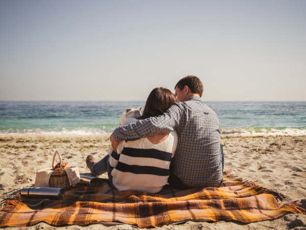A couple sits on a blanket at the beach, looking at the ocean, with a picnic basket nearby under a clear sky.