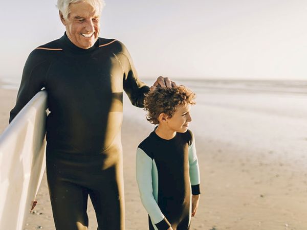 An adult and a child wearing wetsuits stand on the beach with a surfboard, enjoying a sunny day.