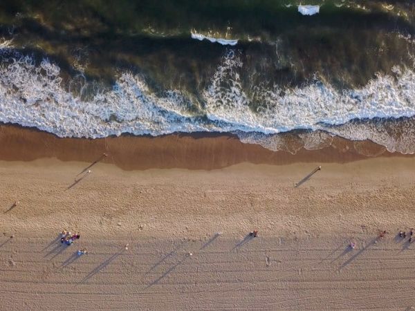 Aerial view of a beach with waves crashing, sand, and people casting long shadows along the shoreline.