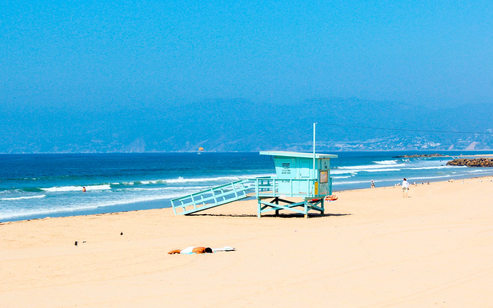 A sandy beach with a lifeguard tower, a few people, and gentle waves under a clear blue sky.