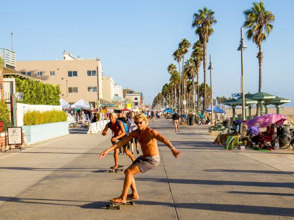 People are skateboarding on a sunny boardwalk lined with palm trees and buildings, creating a lively beachside scene.