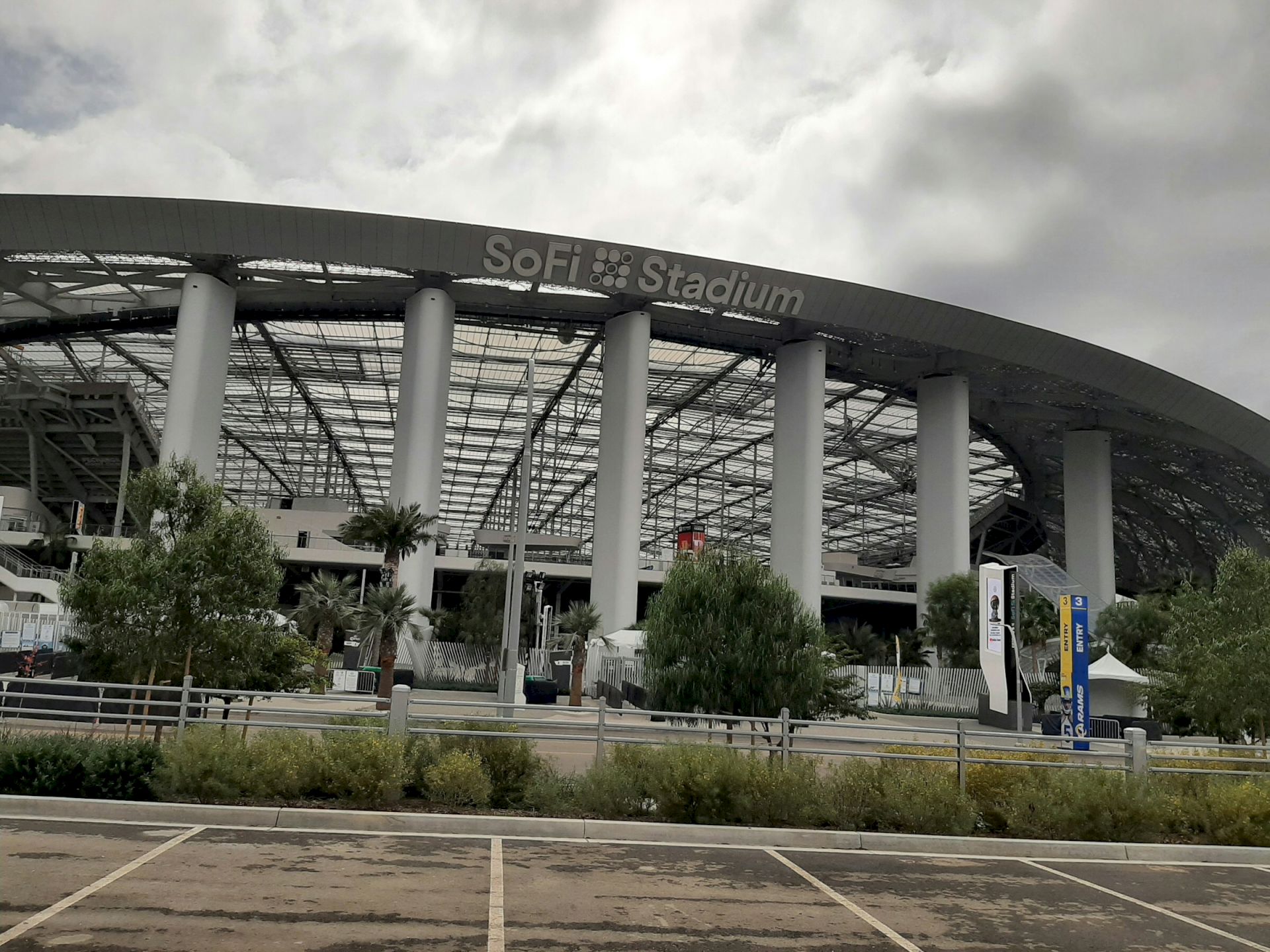The image shows the exterior of a large stadium with "SoFi Stadium" sign, surrounded by trees and a parking area.