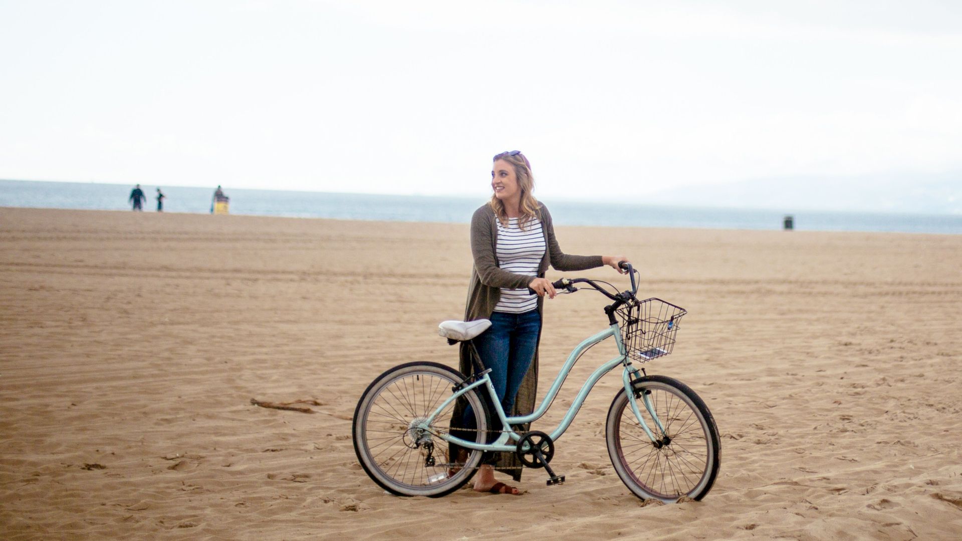 A person stands on a sandy beach with a bicycle, looking at the ocean, with a cloudy sky and distant hills.