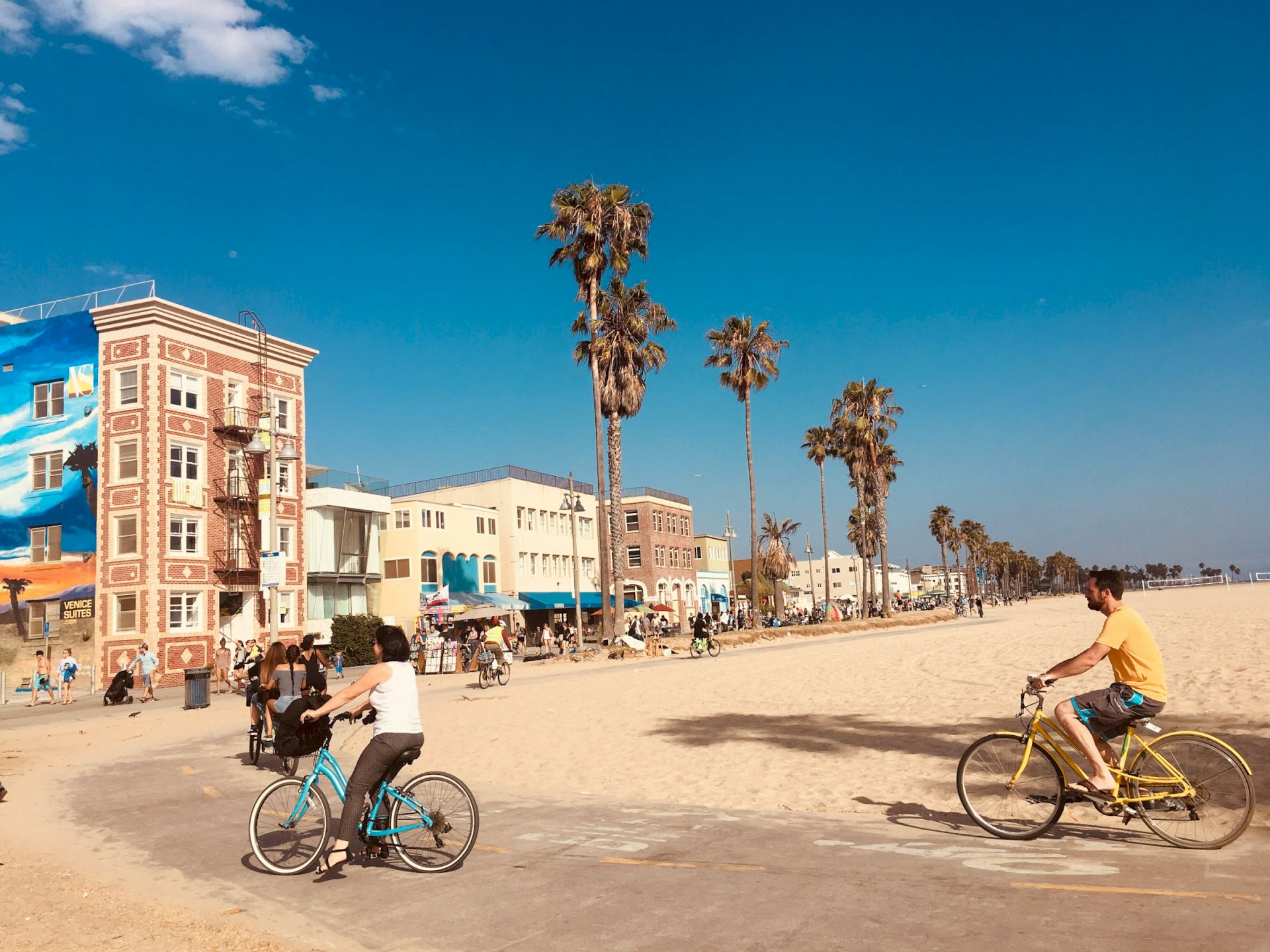People riding bikes along a sunny beach path with palm trees and colorful buildings in the background.