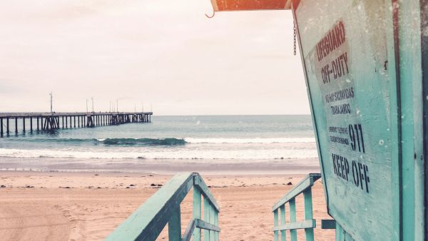 A beach scene with a lifeguard stand in the foreground, a wooden pier, and gentle waves in the background under a cloudy sky.