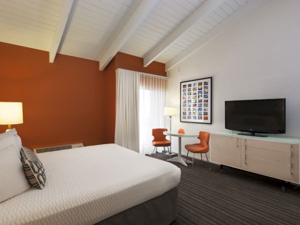 A modern hotel room features a bed, TV, two chairs at a small table, and decorative elements with orange and white accents.