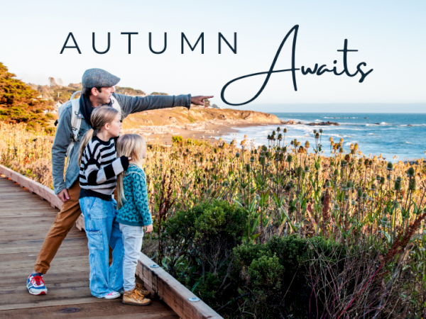 A man and two children look at the ocean on a boardwalk. The text "Autumn Awaits" is written above them.