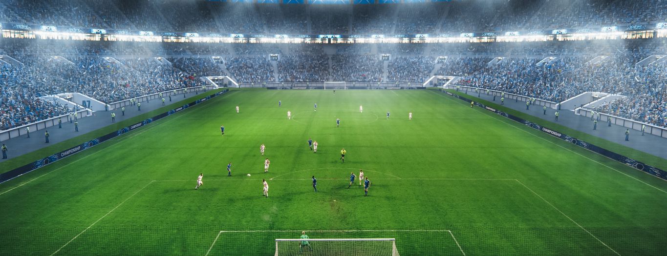 A soccer stadium filled with players on a green field during a match, with bright stadium lights overhead and cheering stands.
