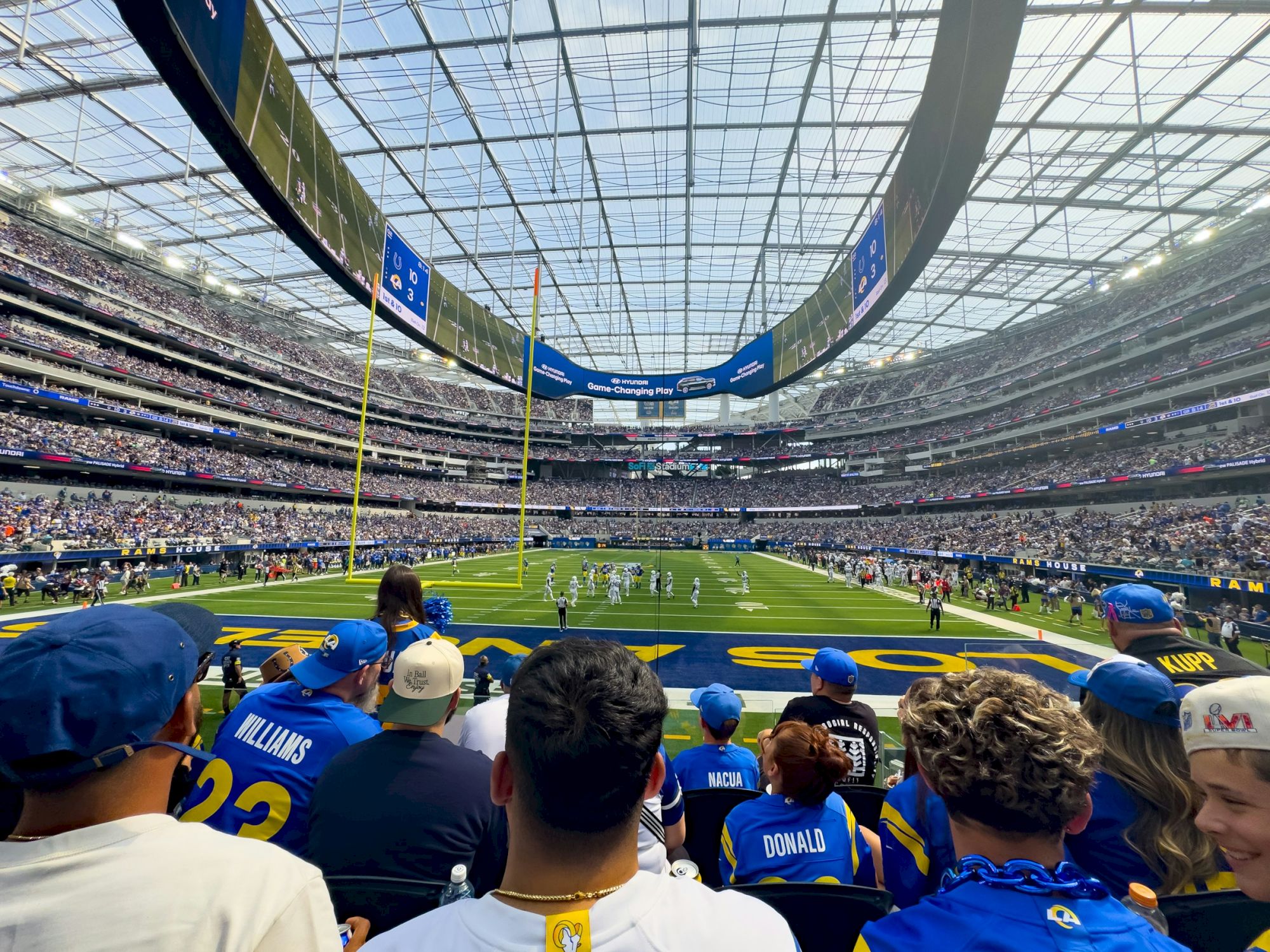 A packed football stadium with fans wearing blue jerseys, watching a game on the bright green field under a glass roof.