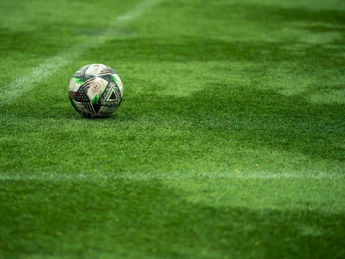 A single soccer ball sits on a lush, freshly mowed green field, drifting near the left side as the grass glistens under soft light.