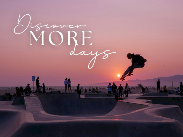 A skater flies off a ramp at a beachside skatepark during sunset, with others watching as the sun sets.