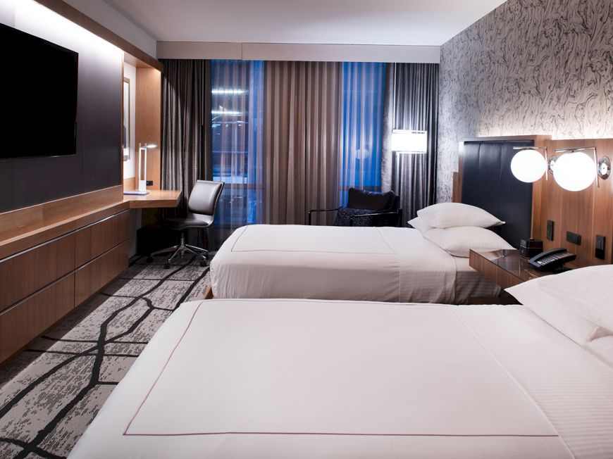 The image shows a modern hotel room with two beds, a TV, and a work desk with a chair, featuring stylish decor and ambient lighting.