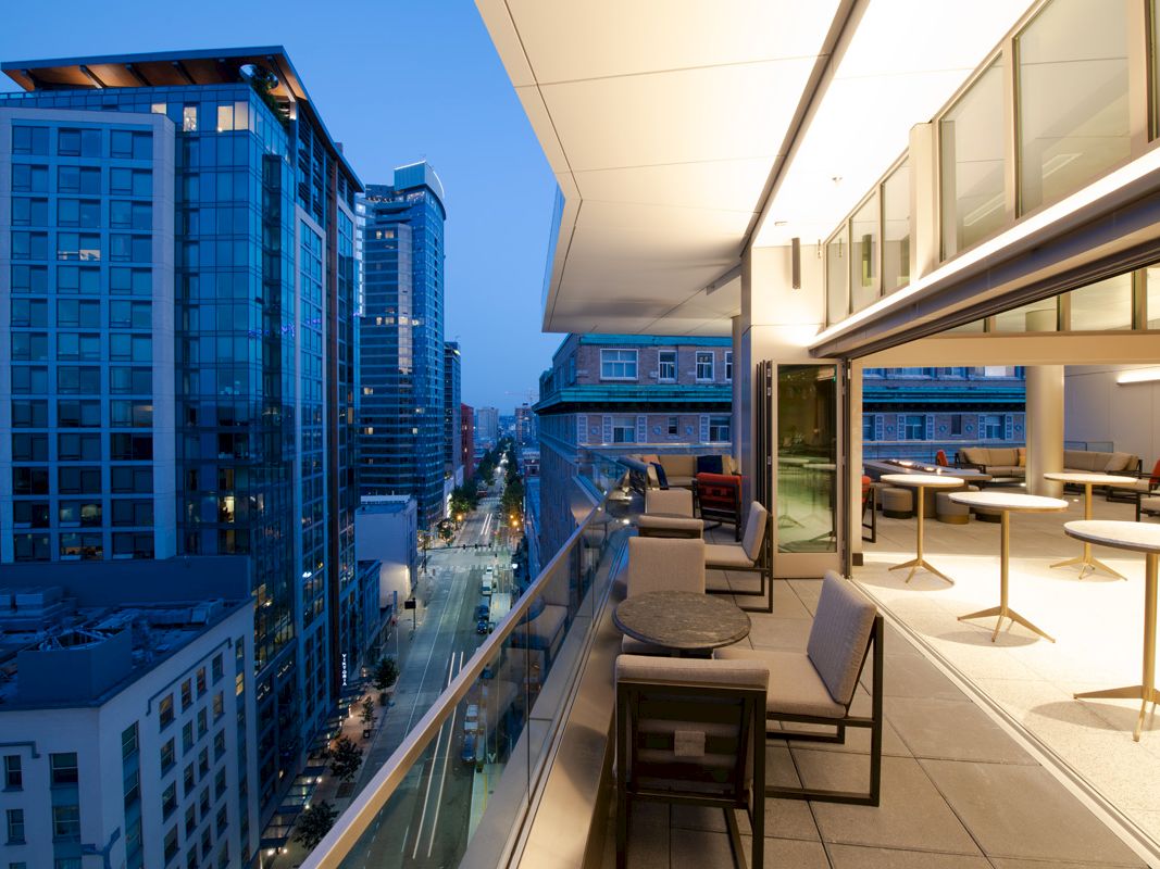 A modern urban balcony view at dusk, overlooking city buildings and a lively street, with outdoor furniture and warm lighting.