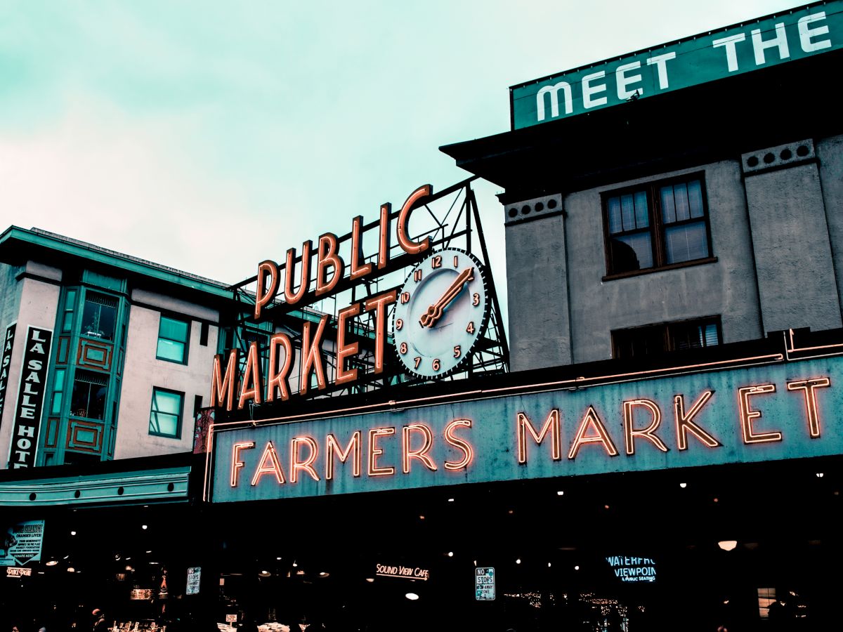 The image shows a public market and farmers market sign with a clock on it, set against an urban backdrop with buildings.