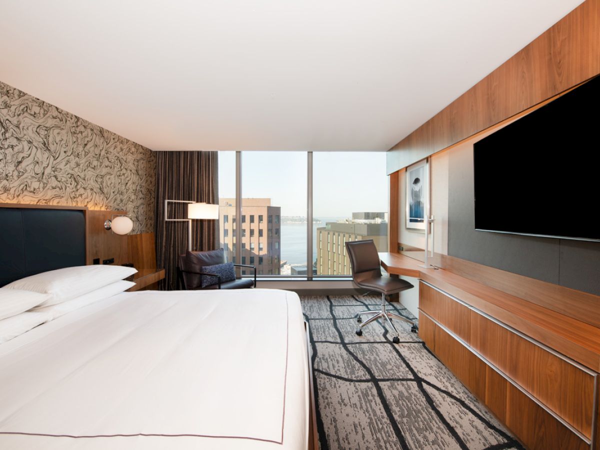 The image shows a modern hotel room with a large bed, desk, chair, wall-mounted TV, and a window view of buildings and water.
