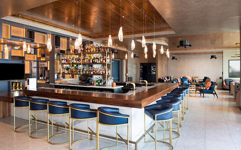 The image shows a modern bar with a wooden counter, blue stools, and pendant lights. Shelves with bottles and a lounge area are visible.
