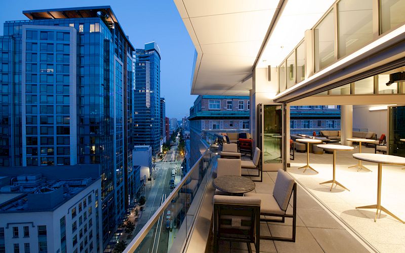 A modern balcony overlooking a cityscape at dusk, featuring outdoor seating and illuminated buildings in the background.