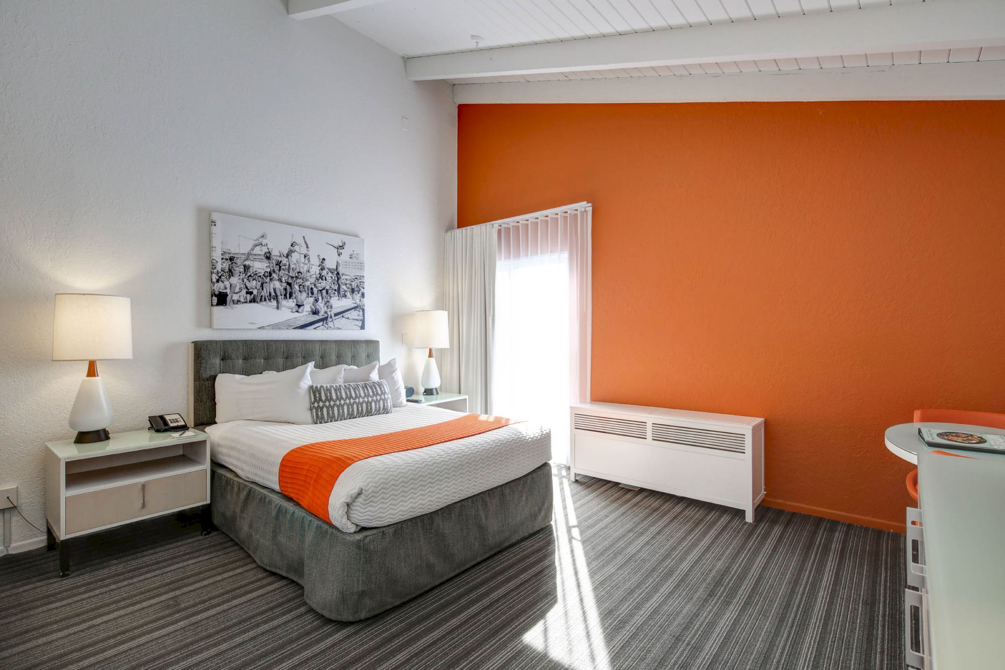 A modern hotel room with orange accents, featuring a bed, lamps, artwork, and a bright window, creating a cozy atmosphere.