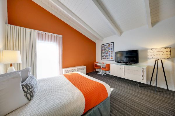 A modern bedroom with an orange accent wall features a bed, TV, desk, chair, and floor lamp, under a vaulted ceiling.