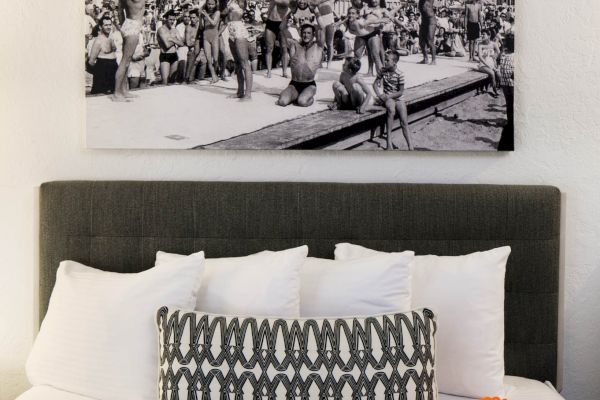 A neatly made bed with white and patterned pillows, an orange object, and a black-and-white photo of people diving by the beach above.