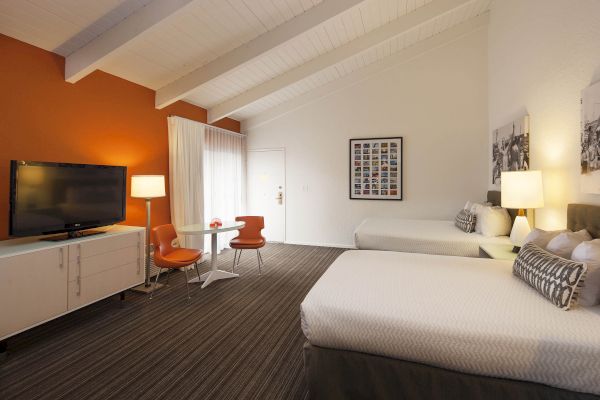 The image shows a hotel room with two beds, a TV, a small table with chairs, and modern decor.
