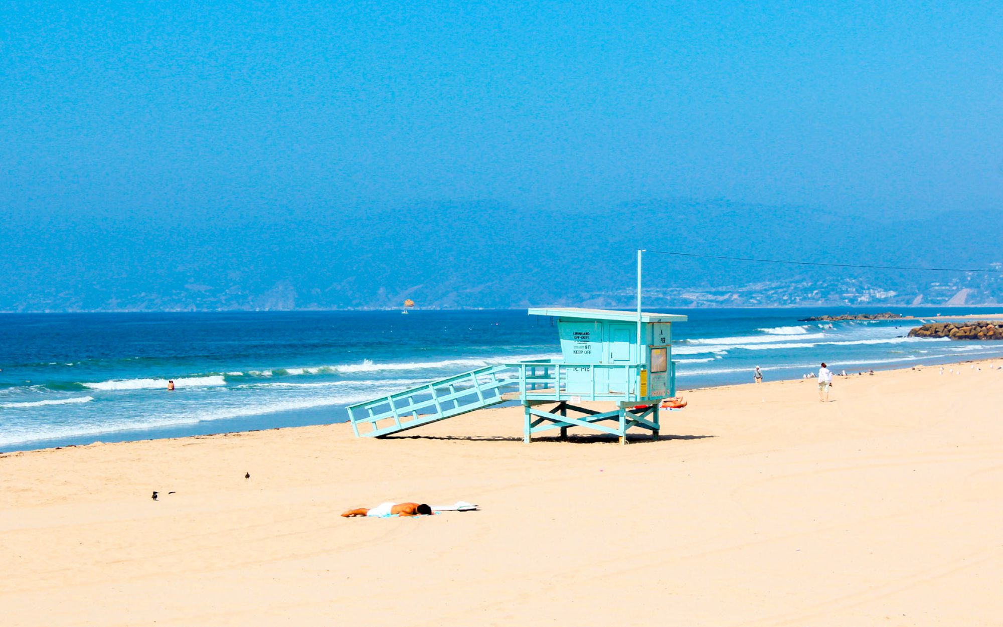 A sandy beach with a lifeguard tower, a few people, and gentle waves under a clear blue sky.