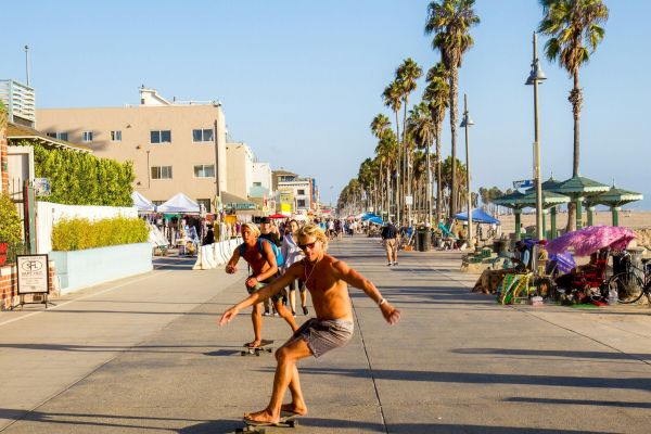 People are skateboarding on a sunny boardwalk lined with palm trees and buildings, creating a lively beachside scene.