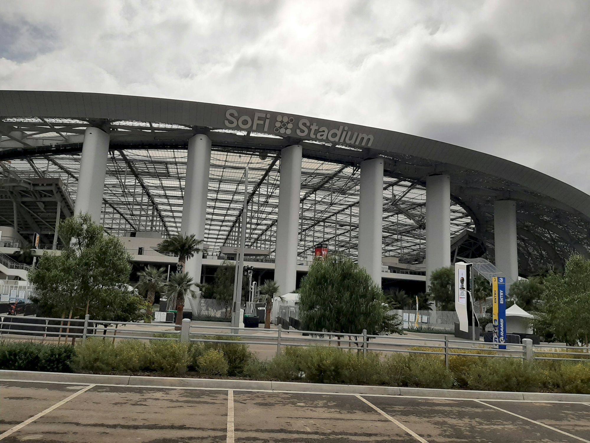 The image shows the exterior of a large stadium with "SoFi Stadium" sign, surrounded by trees and a parking area.