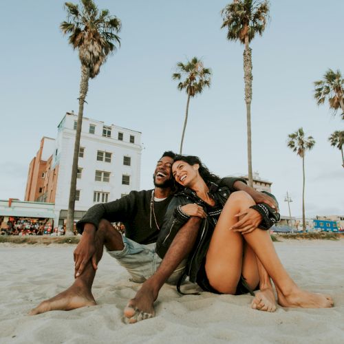 A couple sits on a sandy beach, laughing together with palm trees and a building in the background, under a clear sky.