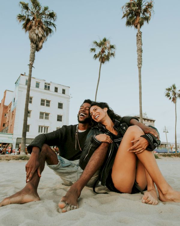 A couple sits on a sandy beach, laughing together with palm trees and a building in the background, under a clear sky.
