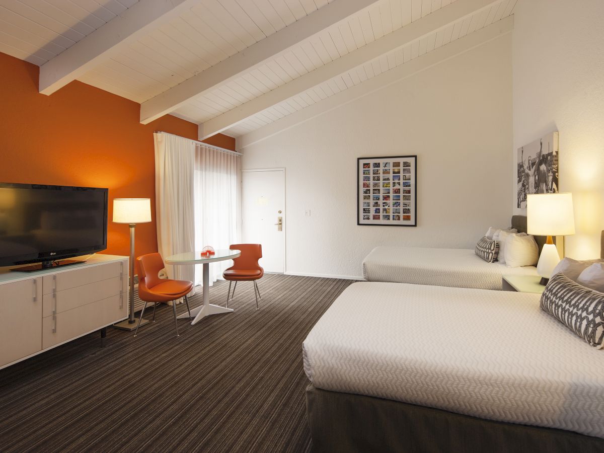 A hotel room with two beds, orange and white decor, a TV, a small table with chairs, a lamp, and framed art.