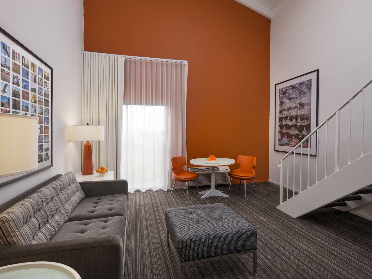 A modern living room with an orange accent wall, gray sofa, orange chairs, a small table, and wall art. Stairs lead to a second level.