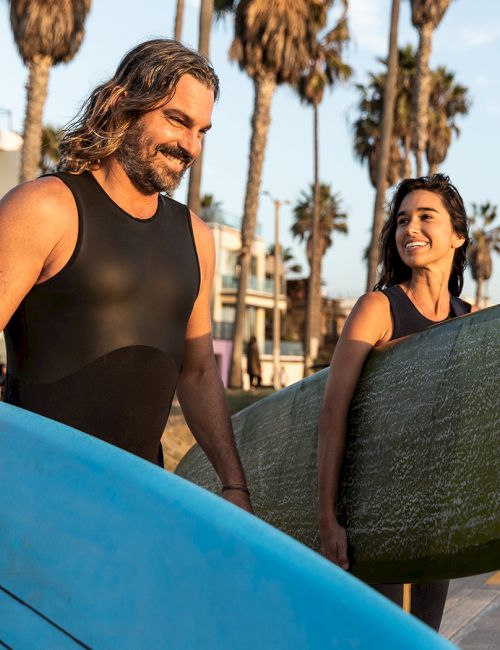 Two people walking on a beach with surfboards, smiling, with palm trees and buildings in the background.