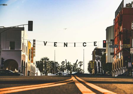 A street scene in Venice, California, with a sign spelling "VENICE" and palm trees, buildings, and cars below a clear sky.
